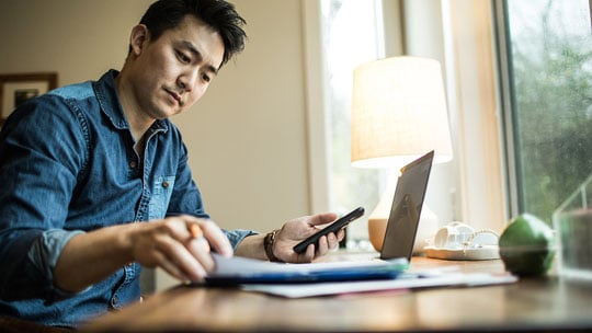 Man sits at desk by window with laptop open and calculator in hand.